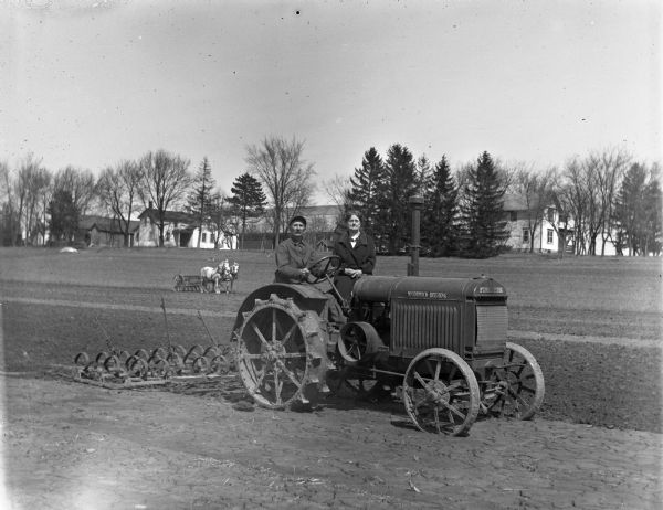 Alexander and Florentina Krueger posing together on McCormick-Deering 10-20 tractor that is pulling a cultivator in a field. A team of two horses pulls another cultivator further down the field. Farmhouses and farm buildings stand along the perimeter of the field.