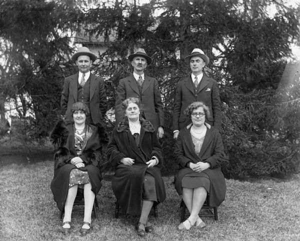 Outdoor group portrait of the Alexander Krueger family. The women are sitting in front on chairs, while the men are standing behind. Front row from the left: Jennie Krueger Bruetzman, Florentina and Elna Krueger, back row: Ernst Bruetzman, Alexander and Edgar Krueger.