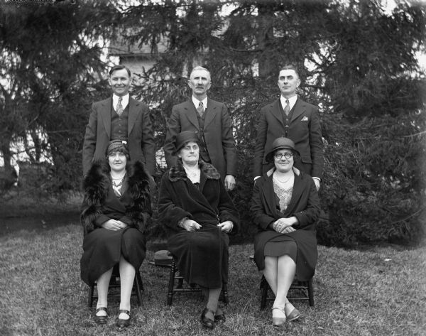 Outdoor group portrait of the Alexander Krueger family. The women are sitting on chairs in the front row, while their husbands are standing behind them. Front row from the left: Jennie Krueger Bruetzman, Florentina, and Elna Krueger. Back row from left: Ernst Bruetzman, Alexander and Edgar Krueger.