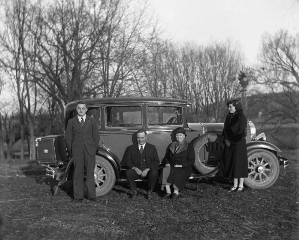 The Bhend family posing with a Studebaker. John Bhend and Sarah Krueger Bhend are sitting together on the running board, while their children, Marcel and Irene, are standing on either side of them. A large trunk is attached to the back of the vehicle.