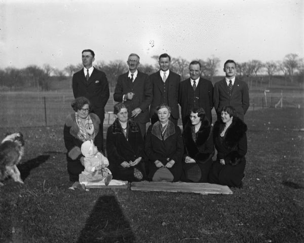 Outdoor group portrait of the Kruegers, Bruetzmans, and Bhends on the Krueger Farm. The women are kneeling on the ground, while the men are standing behind them. The infant, Shirley Krueger, is sitting in front of her mother, Edna Krueger, and a dog is standing on the far left. From left to right: front row, Edna Krueger, Florentina Krueger, Sarah Krueger Bhend, Irene Bhend, and Jennie Krueger Bruetzman; back row, Edgar Krueger, Alexander Krueger, Ernst Bruetzman, John Bhend, and Marcel Bhend.