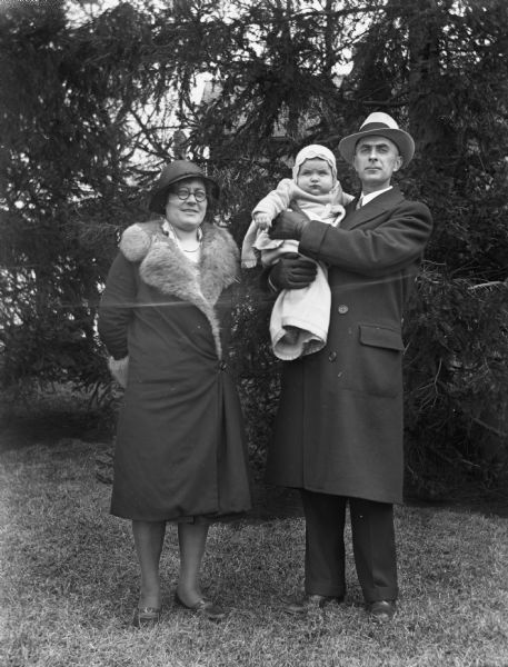 Outdoor group portrait of the Edgar Krueger Family. Elna Krueger stands next to Edgar, who is holding their daughter, Shirley, in his arms.