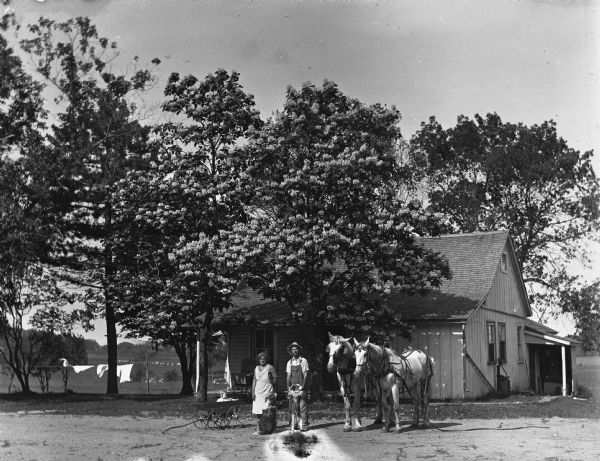 Edgar and Elna Krueger posing in front of an old farmhouse, with a team of two horses and two dogs. Edgar is holding up one of the dogs on its hind legs. Their daughter, Shirley, sits in a small wagon next to Elna.