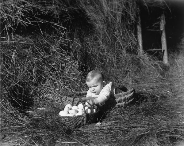 Shirley Krueger sitting in a hand woven basket placed amongst a large pile of hay. She is playing with a stack of eggs in another hand woven basket which is sitting next to her.
