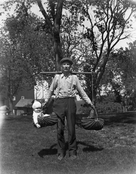 Alexander Krueger carrying Shirley Krueger in a hand woven basket supported by a neck yoke. A barn is in the background.
