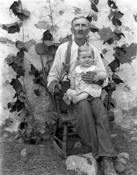 Outdoor portrait of Alexander Krueger sitting in a rocking chair with his granddaughter, Shirley Krueger, sitting in his lap. Ivy is draped on the rocking chair and the building behind it.