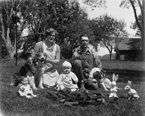 Outdoor portrait of the Edgar Krueger family. Elna and Edgar are kneeling in the grass, each holding a dog at their side, while their daughter, Shirley Krueger, is sitting in between them. Pine cones, a doll, and two bunny rabbit toys are sitting on the ground in front of them.