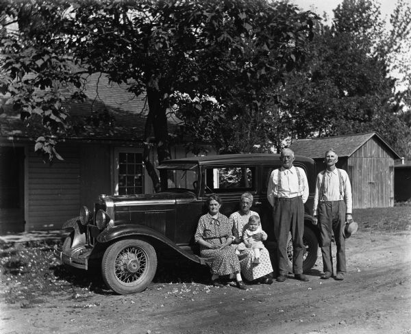 The Kruegers and Sommerfelds posing next to a 1930 Chevrolet in a driveway. Florentina Krueger and Bertha Sommerfeld are sitting along the vehicles running boards with Shirley Krueger sitting in Bertha's lap. William Sommerfeld and Alexander Krueger are standing next to them.