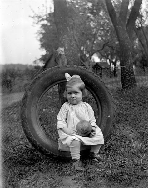 Shirley Krueger sitting in a tire that is propped up against a fence. She is holding a ball in her lap.
