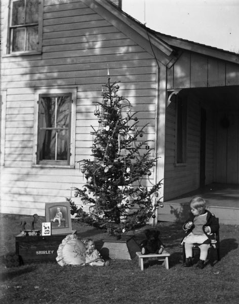 Shirley Krueger sitting in a chair next to a decorated Christmas tree in front of the porch. Several of her toys are placed around the base of the tree.