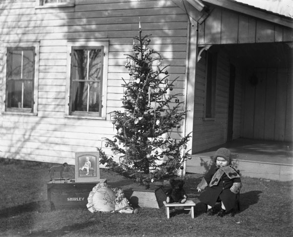 Shirley Krueger sitting a chair next to a decorated Christmas tree that has been placed just off the porch. Several of her presents are around the base of the tree.