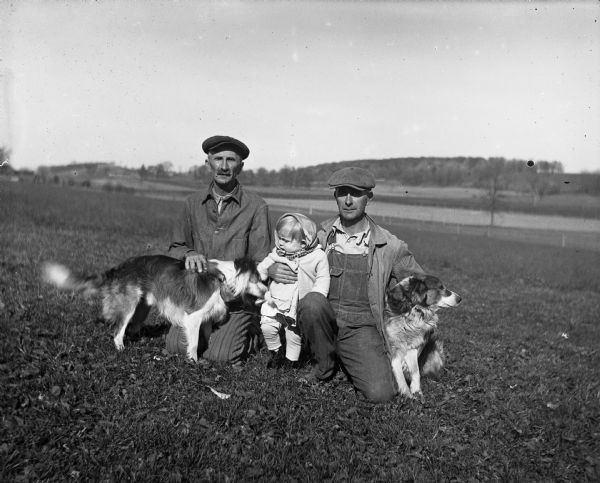 Alexander and Edgar Krueger kneeling in the grass posing with two dogs. Shirley Krueger stands between the two men and is supported by her father, Edgar.