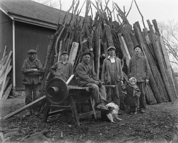 Edgar, Alexander, and Shirley Krueger posing with three other men next to a table saw and an upright pile of stacked wood. A dog is sitting on the ground near the man in the center. Otto Bauman is sitting on the table saw while Frank Tietz and Gilbert Lhettow are standing behind it on the left. Edgar is standing to the right of the saw holding his daughter Shirley's hand, and Alexander is standing on the far right.