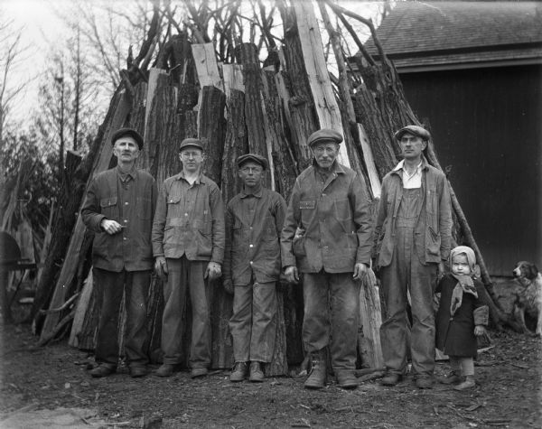 Group portrait of men, with a child, Shirley Krueger, posing in front of an upright pile of stacked wood they are cutting. A dog is sitting on the ground on the right. From left to right: Alexander Krueger, Gilbert Lhettow, Otto Bauman, Frank Tietz, Edgar Krueger, and Shirley Krueger, who is holding her father's hand. In the background is a barn or shed.