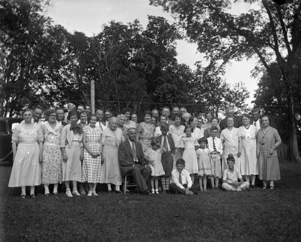 Outdoor group portrait of the entire Krueger family during a family reunion. Henry Krueger sits in a chair in front. Sarah Krueger Bhend and her husband, John Bhend stand on the far left. Next to them is Elna Krueger. Edgar Krueger stands in two back from Elna, holding his daughter, Shirley Krueger. Ernst Bruetzman stands behind Edgar while his wife, Jennie Krueger Bruetzman, stands behind Henry Krueger to the right. Florentina Krueger stands second from the left.