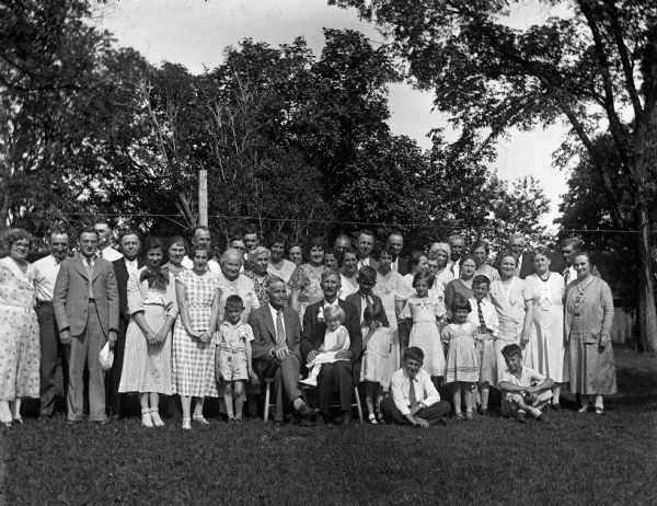 Outdoor group portrait of the entire Krueger family during a family reunion. Henry and Alexander Krueger sit on chairs in front. Shirley Krueger sits on her grandfather's (Alexander's) lap. Jennie Krueger Bruetzman stands behind Alexander while Elna Krueger, John Bhend, and Edgar Krueger stand on the far left. Florentina Krueger stands second in on the far right.