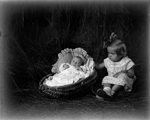 Indoor portrait of Robert and Shirley Krueger sitting in the hay in the barn. Robert is lying in a handwoven basket against a pillow, while his sister is sitting next to him with her arm around the basket.