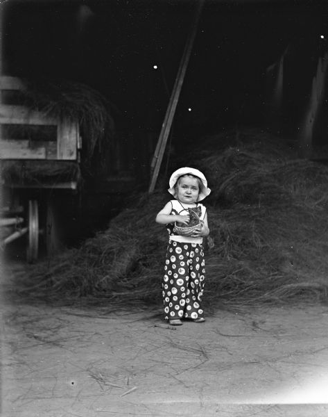Shirley Krueger standing in the hay barn holding a kitten sitting in a small basket. There is a wagon full of hay in the background on the left.