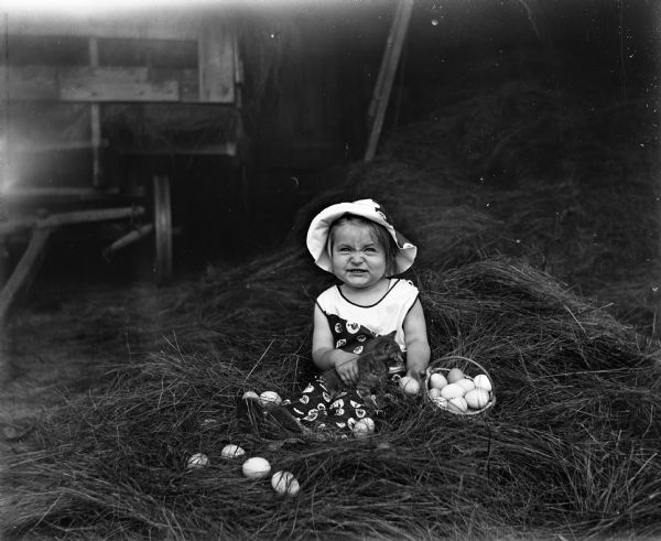 Shirley Krueger sitting in a pile of hay with a kitten in her lap. A basket of eggs is next to her. Several eggs are scattered around her in the hay. In the background on the left is a wagon loaded with hay.
