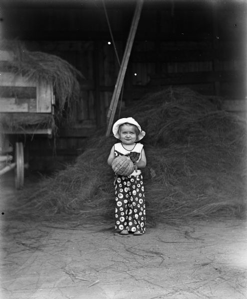 Shirley Krueger standing in the hay barn holding a kitten in a basket. In the background on the left is a wagon full of hay.