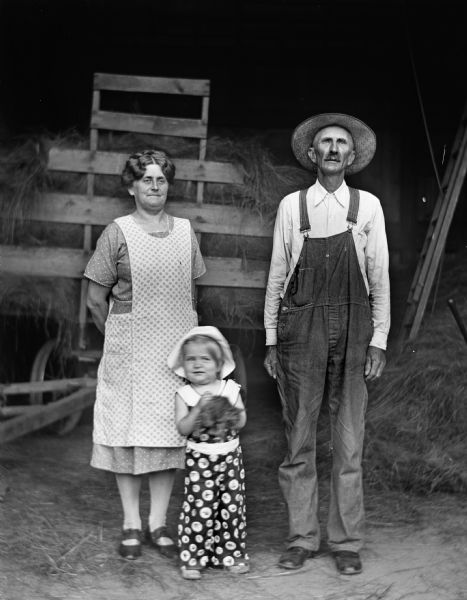 Florentina and Alexander Krueger standing in the hay barn with their granddaughter, Shirley Krueger, who is holding a kitten. Behind them is a wagon loaded with hay.