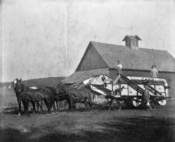 A four-horse team attached to a Case company threshing machine in front of a barn. Edgar and Florentina Krueger are standing on top of the machine, and Alexander Krueger is standing in front with a dog.