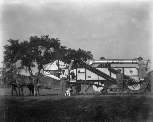 Edgar, Alexander, and Florentina Krueger posing with a Case company threshing machine. Edgar and Alexander are standing in front of the machine, while Florentina is standing in the back on the machine. A team of horses is attached to the machine. A dog is standing next to Edgar.