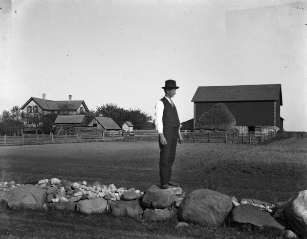 Gust Wendorf standing on a small rock wall. A view of his farm is in the background.