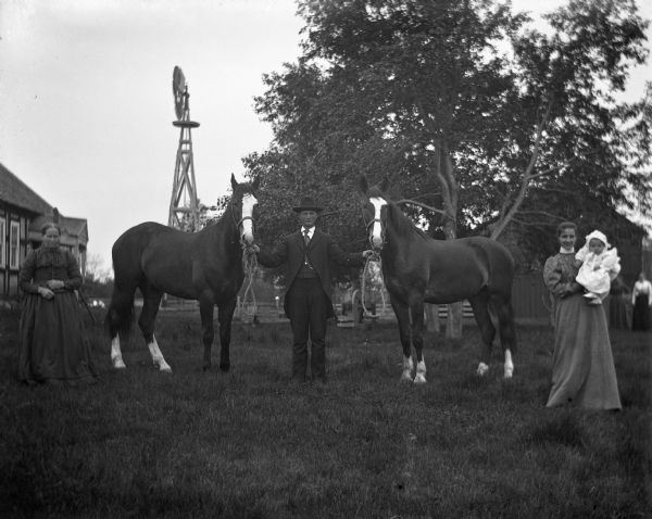 Gust Wendorf, standing in the center, is holding the halters of two horses while his wife, on the right, holds a baby. Mrs. Will stands on the far left, and behind her is what may be a farmhouse, and a windmill. In the background on the right, two women stand next to a farm building.