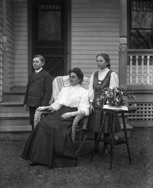 Outdoor group portrait of Florentina Krueger and her children, Edgar and Jennie, in front of their home. Florentina is sitting in a wicker rocking chair while her children are standing on either side of her. An end table with a plant on top is in front of Jennie.