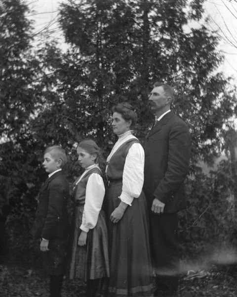 Outdoor group portrait of the Alexander Krueger family in front of a row of pine trees. The family is standing sideways facing left, in a line, ordered from tallest to shortest. From left: Edgar, Jennie, Florentina and Alexander Krueger.