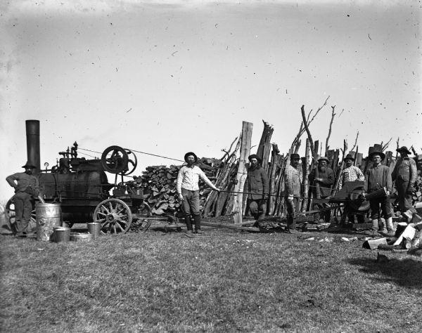 Crew of men sawing wood with the use of a steam engine. A large pile of wood is stacked behind the saw. Alexander Krueger is standing between the steam engine and the belt-driven saw, while August Krueger is standing fourth from the right with five other men. Another man is standing on the far left next to the engine.