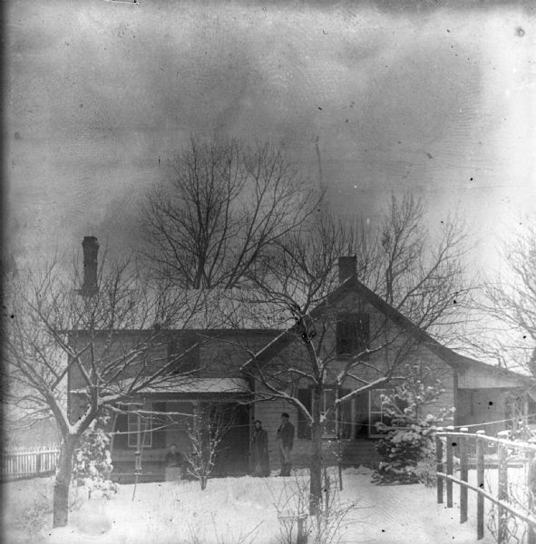 August and Mary Krueger standing in front of their home during winter. Their daughter Sarah Krueger is sitting on the edge of the porch.
