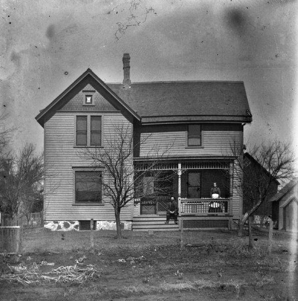 Florentina and Alexander Krueger on the front porch of their home. Alexander is sitting on the porch steps, while Florentina is standing along the railing. Various other farm buildings are behind the home.