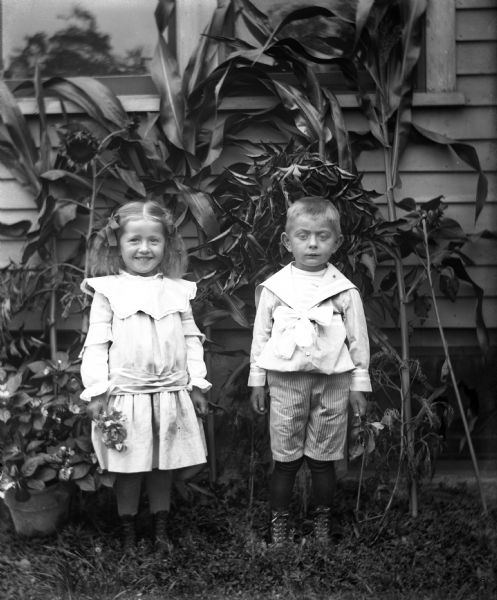 Jennie and Edgar Krueger standing next to the side of the house. They are each holding flowers. Several cornstalks and sunflower plants have been cut and are lying against the house. A potted plant is next to Jennie.