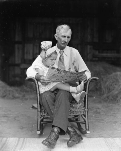 Alexander Krueger sitting outdoors in a wicker rocking chair with his granddaughter, Shirley, sitting in his lap, while he is reading a newspaper.