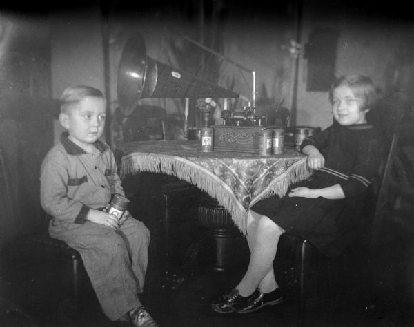 Robert and Shirley Krueger listening to the Edison Standard Phonograph that is sitting on top of the dining room table. Robert is holding one of the cylinder records, and several other records are on the table next to the phonograph.