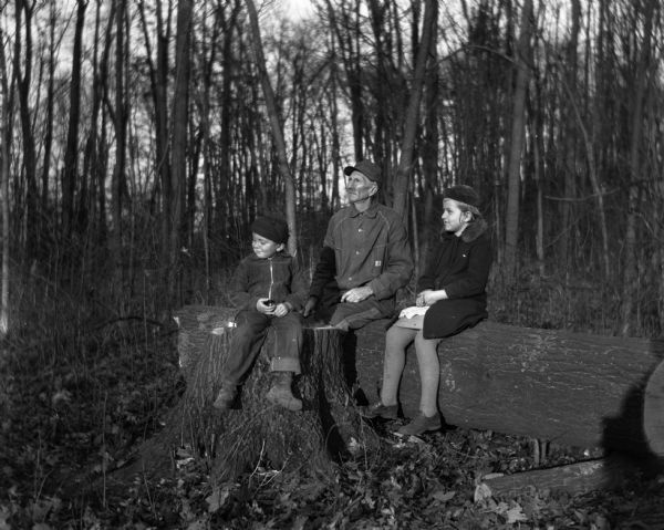 Outdoor portrait of Alexander Krueger sitting on a log with his grandchildren. Shirley Krueger is sitting next to Alexander on the log while Robert is sitting on a stump in front.