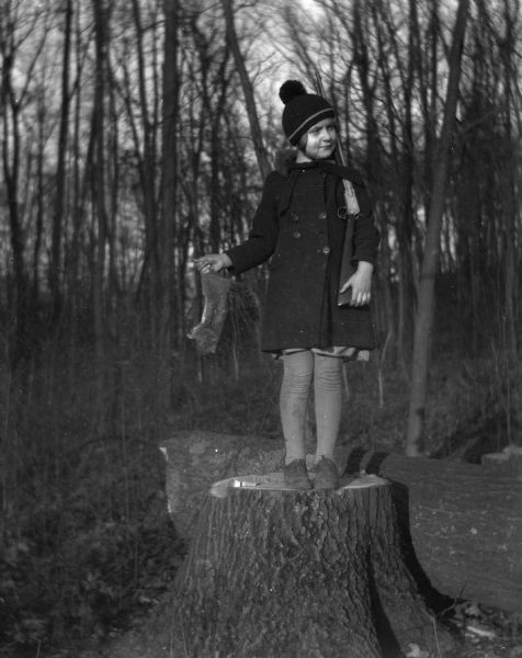 Shirley Krueger standing on a stump holding a dead squirrel in her right hand while propping a rifle on her left shoulder.