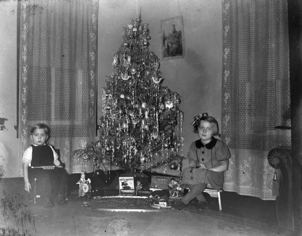Robert and Shirley Krueger sitting in chairs on either side of a decorated Christmas tree. Several of their toys are displayed under the tree. Robert is holding a small trumpet in his lap.