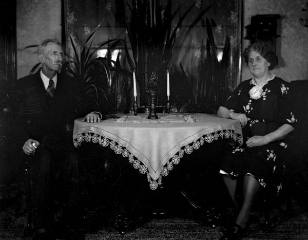 Indoor portrait of Alexander and Florentina Krueger sitting at either end of the dining room table. The table is set with a knit table cloth and candelabra. Several plants are behind the table.