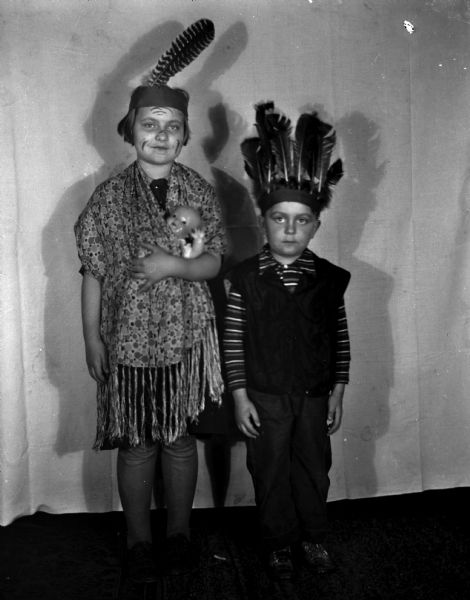 Shirley and Robert Krueger dressed up as Native Americans and wearing feather headdresses. Shirley is wearing a shawl with a baby doll swaddled in between, and face makeup. A white cloth is hanging behind them as a backdrop.