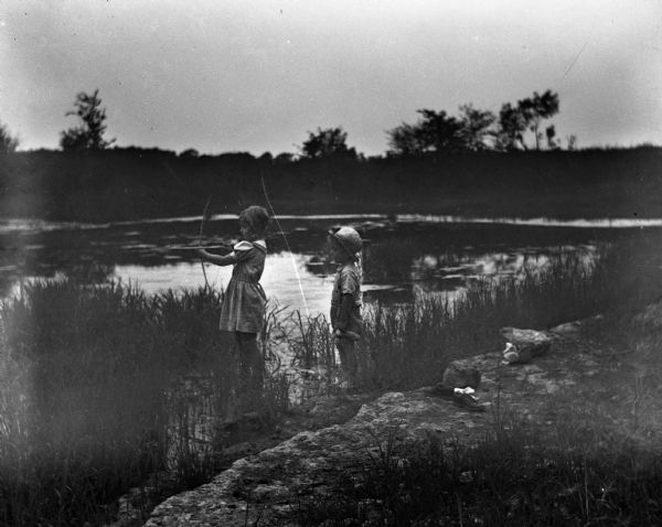 Shirley and Robert Krueger standing barefoot at the edge of a pond. Their shoes and socks are lying on rocks at the waters edge. Shirley is holding a bow with an arrow ready to shoot while Robert is standing behind her.