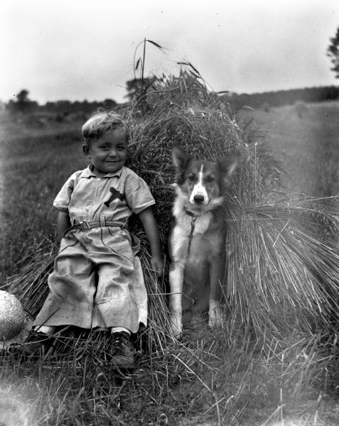 Outdoor portrait of Robert Krueger sitting on a grain shock with his dog, Sport. His straw hat is lying on the ground next to his feet.