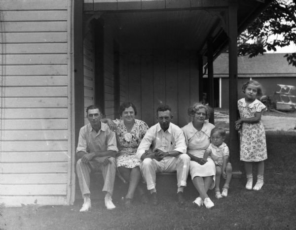 The Edgar Krueger family sitting on the edge of the porch with friends. From left to right: Edgar Krueger, Ann Buss, Adolf Buss, Elna Krueger, and Robert Krueger. Shirley is standing, leaning against the post supporting the porch.