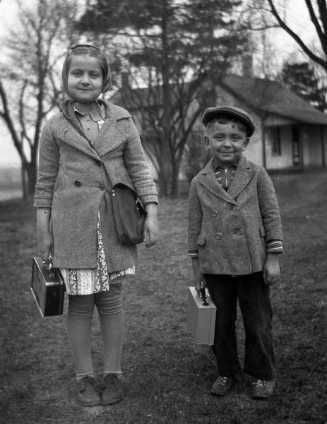 Outdoor portrait of Shirley and Robert Krueger standing in the front lawn, ready for school. Each of them are carrying a small briefcase for school, while Shirley is carrying a small messenger bag.