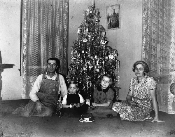 The Edgar Krueger family sitting on the floor in front of the Christmas tree. From left to right: Edgar, Robert, Shirley, and Elna Krueger. Several of the children's new toys are near them on the floor.