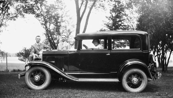Elna Krueger sitting in the driver's seat of a Chevrolet. Edgar Krueger is standing on the other side of the car, near the hood, while a friend is standing in front resting his foot on the bumper.