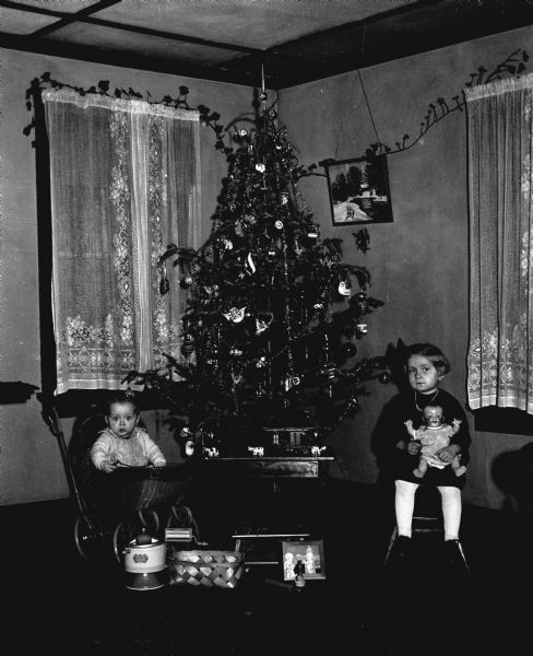 Shirley and Robert Krueger sitting next to a decorated Christmas tree. Robert is sitting in a baby carriage while Shirley is sitting in a chair. Several of their Christmas presents are placed in front of the tree.
