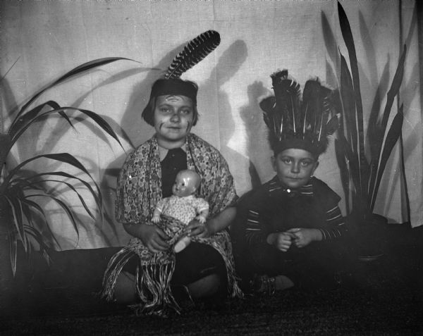 Shirley and Robert Krueger sitting cross legged on the floor wearing feather headdresses, dressing as Native American Indians. Shirley is wearing face makeup and a shawl while holding a baby doll. They are sitting in front of a white cloth backdrop with two potted plants on either side of them.